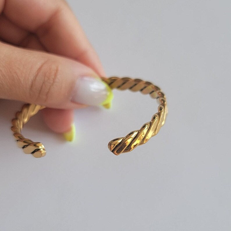 Gold twisted ring held by a hand with light nail polish on a plain background